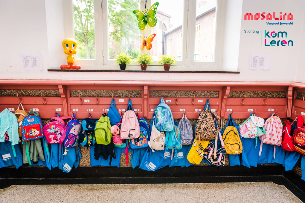 Garderobe in een basisschool met kleurrijke kinderrugzakken, jasjes en gymtassen aan haakjes onder een raam, met het logo van MosaLira en Stichting Kom Leren op de muur.